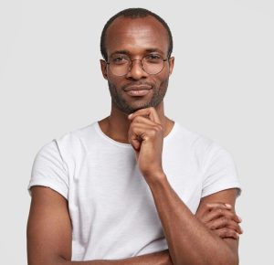 Studio shot of serious black man keeps hand under chin, wears transparet glasses and casual t shirt, keeps hands partly crossed, poses against white background, thinks about something, being clever